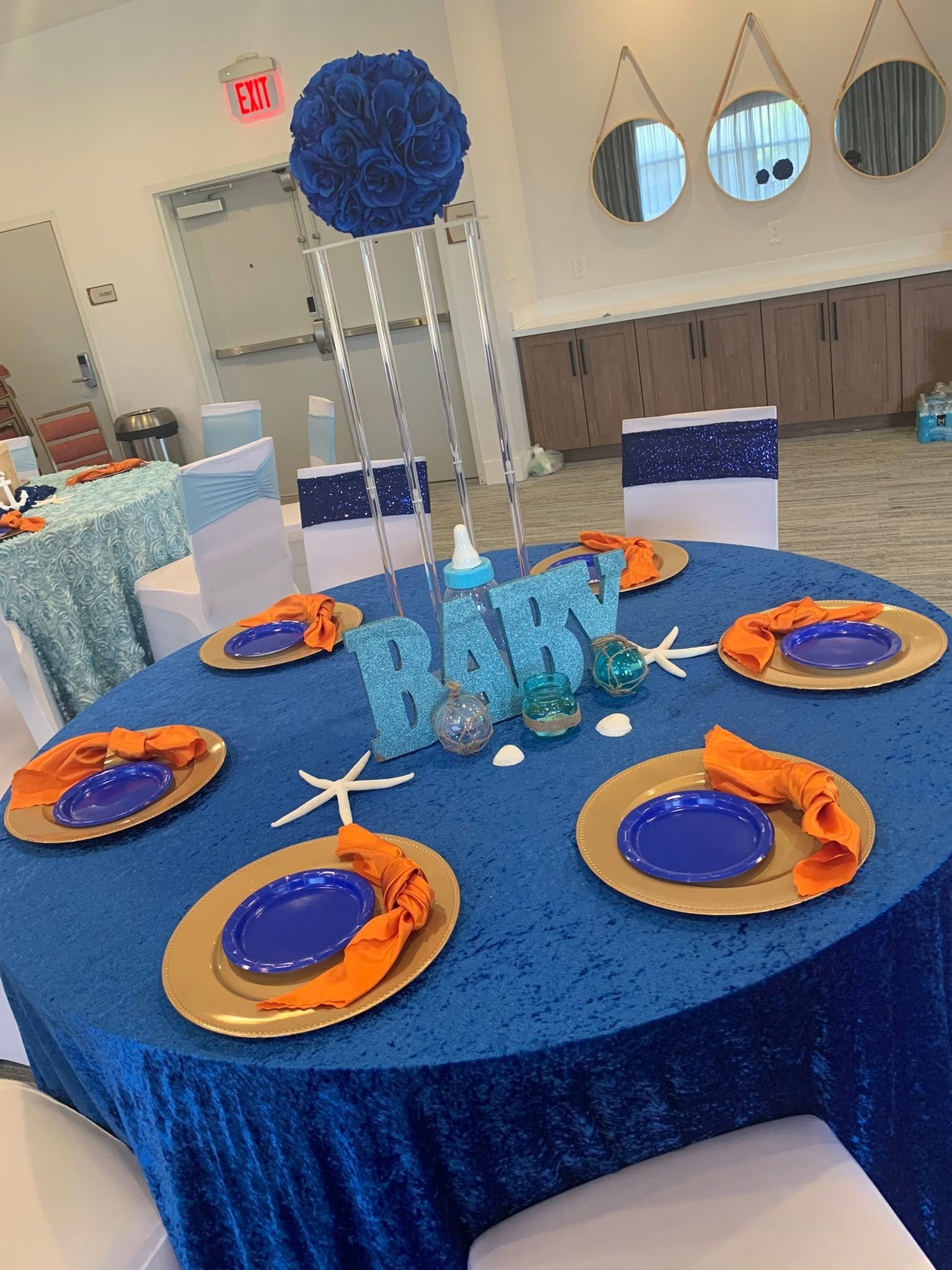 Royal blue baby shower table with gold chargers, orange napkins and a blue rose centerpiece