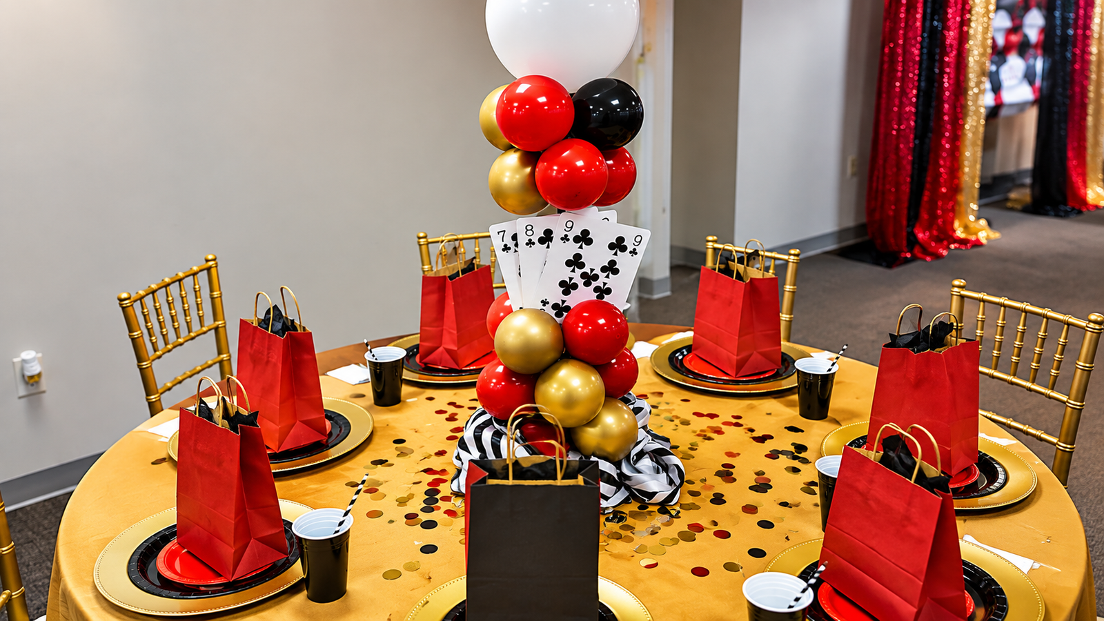 Casino themed table with a red, black and gold balloon centerpiece, playing cards and gold chiavari chairs